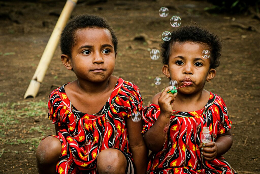 pexels-photo-10404339-10404339 Two young children in vibrant traditional attire blowing bubbles outdoors in Port Moresby.